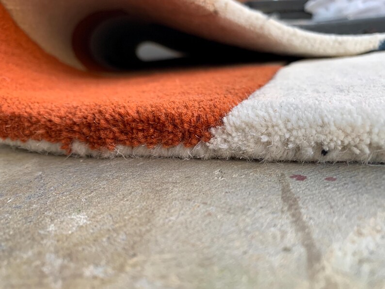 Puede incluir: Primer plano de una alfombra redonda enrollada con una superficie texturizada. La alfombra presenta un dise&ntilde;o de bloques de color con secciones naranjas y blancas. El borde de la alfombra es visible, mostrando el grosor y la textura del material.