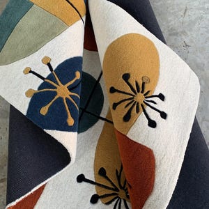 Puede incluir: Una alfombra enrollada con un dise&ntilde;o moderno de mediados de siglo. La alfombra presenta un fondo crema con formas abstractas en azul marino, amarillo mostaza, &oacute;xido y verde oliva. Tambi&eacute;n hay detalles florales negros.