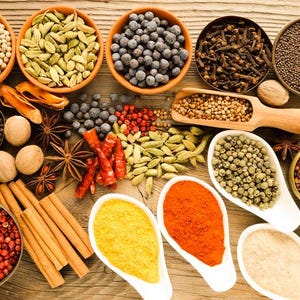 May include: Overhead shot of various spices and herbs in bowls and spoons on a wooden surface. Includes peppercorns, cardamom, cloves, mustard seeds, nutmeg, cinnamon sticks, and chili peppers.