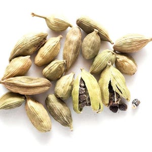 Puede incluir: Un conjunto de vainas de cardamomo verdes y beige, algunas abiertas para revelar semillas oscuras, sobre un fondo blanco. Las vainas tienen una superficie texturizada y están dispuestas de forma dispersa.