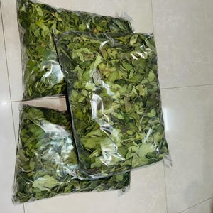 Può includere: Tre sacchetti di plastica trasparente riempiti con foglie verdi essiccate. I sacchetti sono rettangolari e contengono una grande quantità di fogliame disidratato, che mostra varie sfumature di verde e alcuni bordi marroni, compattati.