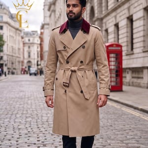 Op de afbeelding: Een beige dubbelbrede trenchcoat met een bordeauxrode kraag en riem. De jas heeft een klassiek ontwerp met een ceintuur in de taille en knopen. De achtergrond toont een geplaveide straat en gebouwen.