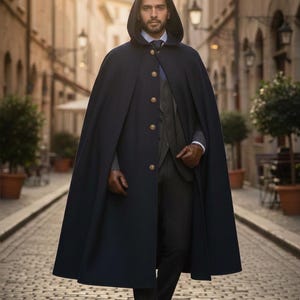 May include: A dark navy blue hooded cape featuring a row of gold-toned buttons down the front. The garment is worn over a dark grey suit jacket, a light blue collared shirt with a dark tie, and dark trousers, paired with brown leather gloves and shoes.