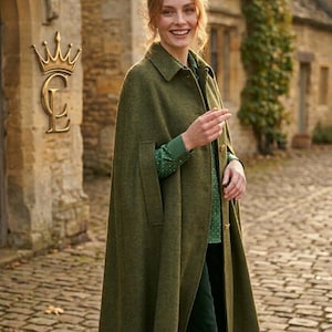 May include: An olive green wool-blend cape coat featuring a classic collar, a button-front closure, and arm slits. The garment is worn over a green long-sleeved shirt with a subtle white pattern and dark green trousers, paired with brown boots.