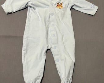 May include: Light blue baby sleeper with long sleeves and attached feet. The sleeper has a button-up front and a small embroidered design of cartoon characters. A tag is attached near the collar.