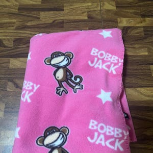 May include: A bright pink fleece blanket featuring a repeating pattern of white stars and a cartoon monkey character with a wide grin. The text BOBBY JACK is printed in white lettering throughout the fabric, which has a soft, textured surface.