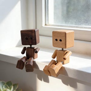 Puede incluir: Dos figuras de bloques de madera con brazos y piernas móviles. Una es de color marrón oscuro, la otra marrón claro. Ambas tienen una cabeza cuadrada con dos pequeños detalles oscuros para los ojos. Están sentadas en un alféizar blanco.