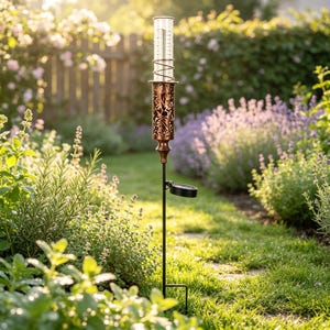 May include: A copper-colored metal rain gauge featuring an intricate cut-out floral pattern on the cylindrical holder. The clear glass tube is marked with measurement lines. A small solar panel is attached to the thin black metal stake.