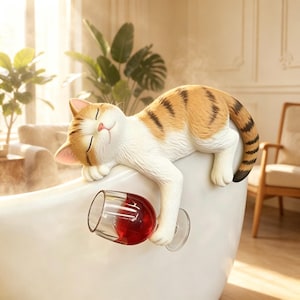Puede incluir: Una figura caprichosa de un gato durmiente con pelaje naranja y blanco, descansando en el borde de una bañera blanca. La pata del gato sostiene una copa de vino llena de líquido rojo. La escena está ambientada en una habitación luminosa con plantas.
