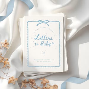 Könnte beinhalten: Ein Stapel Babyparty-Andenkenbücher mit dem Titel "Letters to Baby". Das Cover zeigt hellblauen Text und einen dekorativen Rahmen. Die Bücher liegen auf einer weißen Oberfläche mit blauem Band und Trockenblumen.