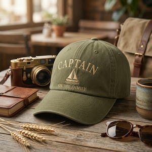 May include: Olive green baseball cap with the word "CAPTAIN" embroidered in gold, along with a sailboat graphic. The text "ON THE FAMILY" is below the sailboat. The hat is on a wooden surface with a vintage camera and sunglasses.
