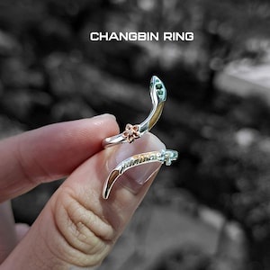 May include: A silver ring with a snake design, featuring a flower and green accents. The ring is wrapped around a finger. The text "CHANGBIN RING" is displayed at the top.