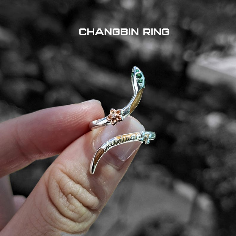May include: A silver ring with a snake design, featuring a flower and green accents. The ring is wrapped around a finger. The text "CHANGBIN RING" is displayed at the top.