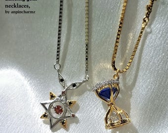 May include: Two necklaces with box chains. The silver-tone necklace features a star-shaped pendant with a central red gem and small black accents. The gold-tone necklace features an hourglass pendant with a blue gem and a clear stone at the base.