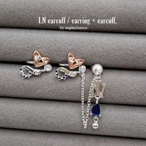 Può includere: Set di polsini e un orecchino argentati su sfondo grigio. I polsini presentano un design a volpe in oro rosa e argento, con dettagli blu e trasparenti. L'orecchino ha una catena, una gemma blu a goccia e le lettere "IN". Il testo "I.N earcuff / earring + earcuff, by anpincharmz" è visibile.