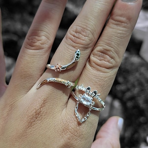 May include: A hand displays four silver rings. One ring is a snake design with green and gold accents. Another ring has a floral design. A third ring has a chain and a crystal. The fourth ring has a leaf design.