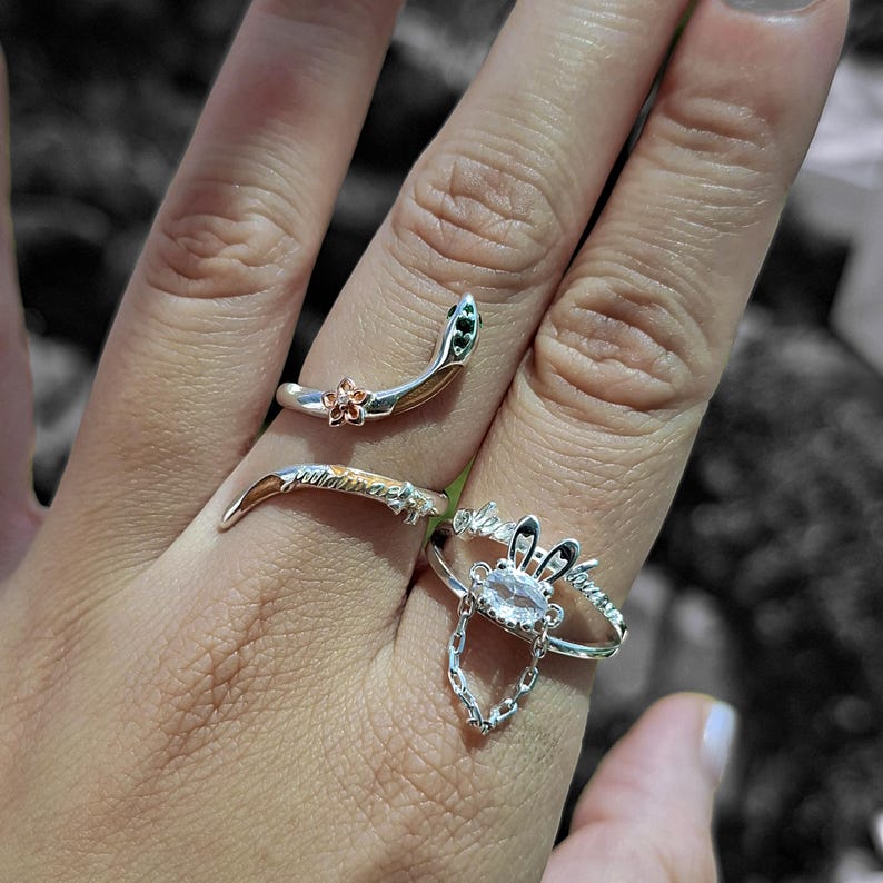 May include: A hand displays four silver rings. One ring is a snake design with green and gold accents. Another ring has a floral design. A third ring has a chain and a crystal. The fourth ring has a leaf design.