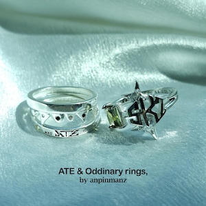 Puede incluir: Dos anillos de plata con diseños únicos. Uno presenta un diseño geométrico con detalles negros y el texto "WHO ATE THE". El otro tiene un diseño de estrella y geométrico con una gema verde y las letras "SKK".