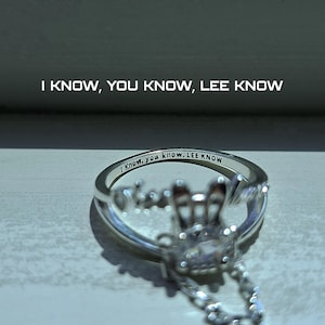 May include: A silver ring with the inscription "I Know, you know, LEE KNOW" engraved on the band. The ring features a decorative design with clear gemstones and the text "I KNOW, YOU KNOW, LEE KNOW" above the ring.