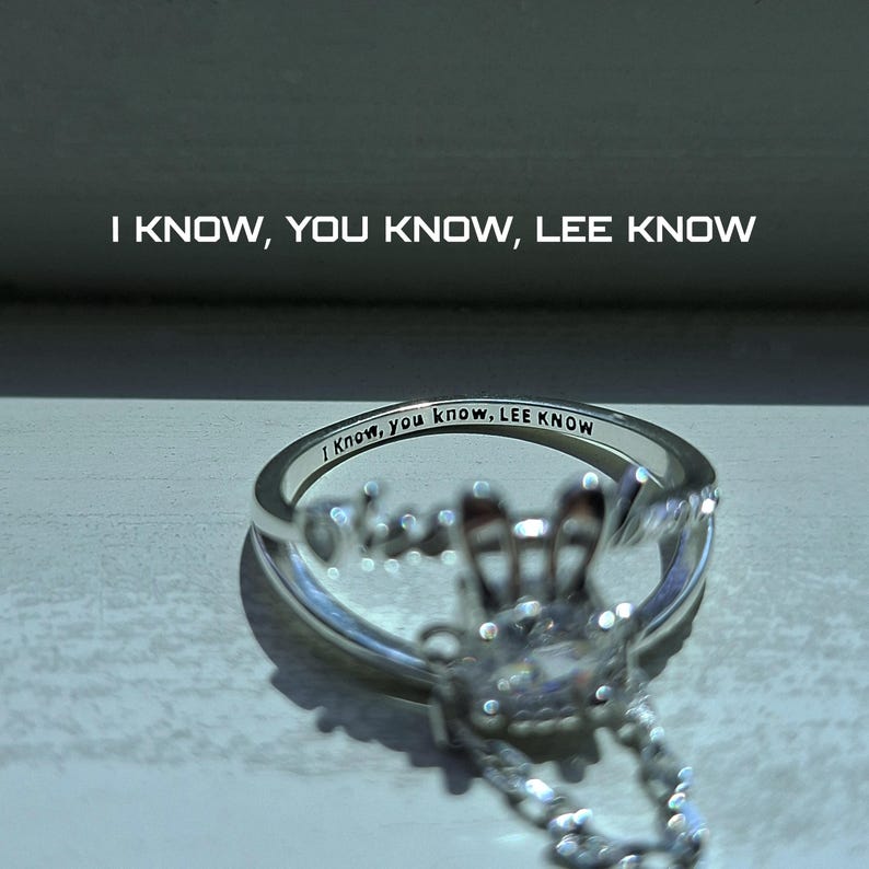 May include: A silver ring with the inscription "I Know, you know, LEE KNOW" engraved on the band. The ring features a decorative design with clear gemstones and the text "I KNOW, YOU KNOW, LEE KNOW" above the ring.
