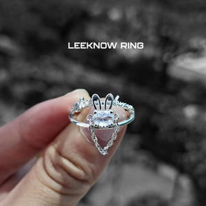 May include: A silver ring featuring a bunny design with a central oval-cut gemstone and dangling chain. The ring has the text "LEEKNOW RING" above it. The ring is held up by a person's hand.