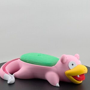 Pu&ograve; includere: Un portasapone rosa a forma di Slowpoke con una saponetta verde. Il portasapone ha un becco giallo, bocca rossa e dettagli bianchi. Il prodotto &egrave; progettato per contenere una saponetta.