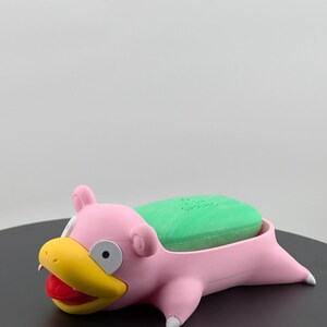 Pu&ograve; includere: Un portasapone rosa a forma di Slowpoke con becco giallo, bocca rossa e dettagli bianchi. Una saponetta verde riposa nel portasapone. Il portasapone &egrave; su una superficie nera.