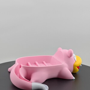 Pu&ograve; includere: Un portasapone rosa a forma di personaggio dei cartoni animati, con becco giallo e lingua rossa. Il portasapone ha un porta sapone rettangolare e una coda curva. Il personaggio &egrave; sdraiato su una superficie nera.
