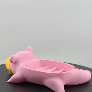 Pu&ograve; includere: Un portasapone rosa a forma di animale dei cartoni animati, forse un maiale o un ippopotamo, con becco giallo e dettagli bianchi. Il portasapone ha un bacino rettangolare con creste.