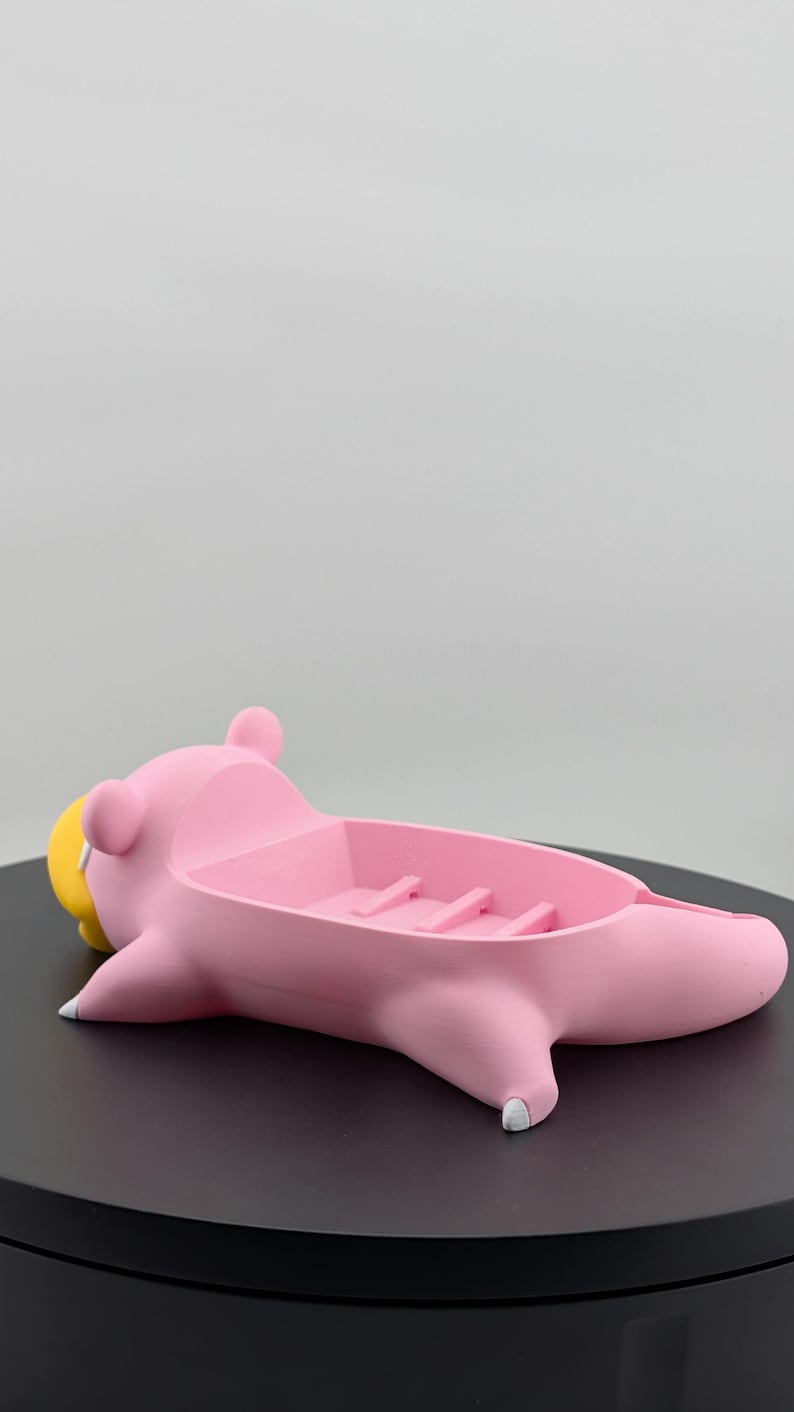 Pu&ograve; includere: Un portasapone rosa a forma di animale dei cartoni animati, forse un maiale o un ippopotamo, con becco giallo e dettagli bianchi. Il portasapone ha un bacino rettangolare con creste.