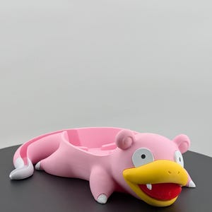 Pu&ograve; includere: Un vassoio rosa a forma di Slowpoke con dettagli bianchi su piedi e coda. Il vassoio ha un becco giallo, bocca rossa e occhi neri. Progettato per contenere piccoli oggetti, &egrave; posto su una superficie nera.