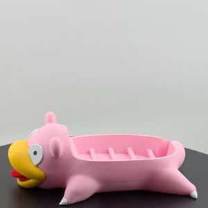 Pu&ograve; includere: Un portasapone rosa a forma di personaggio dei cartoni animati Slowpoke. Ha un becco giallo, una lingua rossa e dettagli bianchi. Il portasapone ha un'area rettangolare con divisori rialzati.