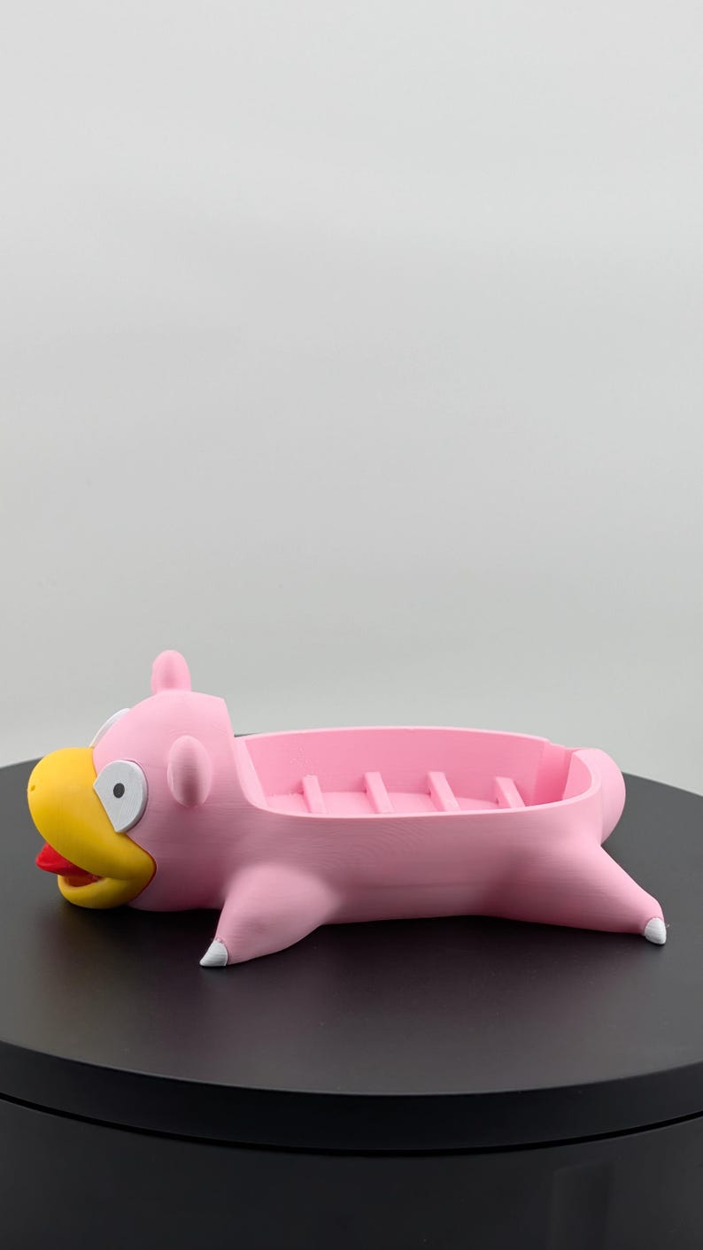 Pu&ograve; includere: Un portasapone rosa a forma di personaggio dei cartoni animati Slowpoke. Ha un becco giallo, una lingua rossa e dettagli bianchi. Il portasapone ha un'area rettangolare con divisori rialzati.