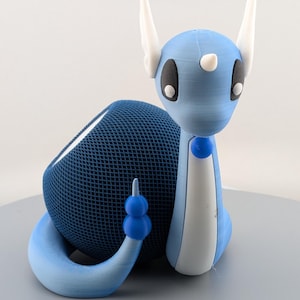 Puede incluir: Una figura de dragón impresa en 3D en azul claro y blanco, con grandes ojos negros, cuernos blancos y una cola azul. El dragón está posado junto a un objeto de malla azul oscuro, posiblemente un altavoz. La figura está sobre una superficie gris.