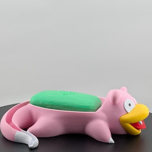Pu&ograve; includere: Un portasapone rosa stampato in 3D a forma di personaggio dei cartoni animati, Slowpoke. Contiene una saponetta verde. Lo Slowpoke ha un becco giallo, una lingua rossa e dettagli bianchi. Il portasapone &egrave; su una superficie nera.