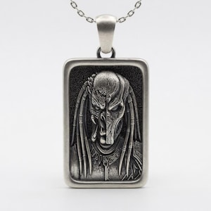 May include: A rectangular silver-toned pendant on a chain featuring a detailed relief of a creature with dreadlocks, showing a split face design with one side revealing a textured alien mask and the other side showing a more organic, humanoid facial structure.