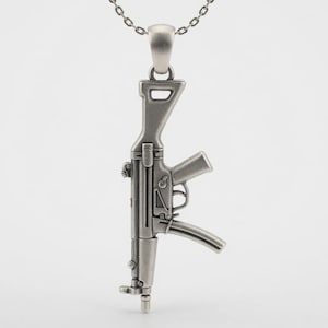May include: A silver-toned metal pendant shaped like a rifle, suspended from a matching silver-toned chain. The pendant features detailed engravings of the firearm's barrel, stock, trigger guard, and magazine, with a matte finish throughout.