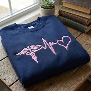 May include: Navy blue sweatshirt with a pink embroidered design featuring a caduceus symbol, a heartbeat line, and a heart. The sweatshirt is folded on a wooden surface, with books and a small plant in the background.