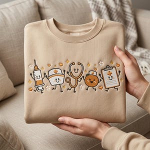 May include: A beige crewneck sweatshirt featuring an embroidered design of five anthropomorphic medical items including a syringe, a nurse cap, a stethoscope, an orange, and a clipboard, all with smiling faces, surrounded by small stars, hearts, and flowers.
