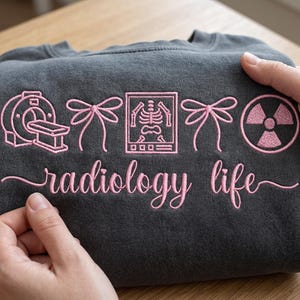 Puede incluir: Sudadera gris oscuro con diseños bordados en rosa con temática de radiología. El diseño incluye un escáner CT, un esqueleto y un símbolo de radiación, con las palabras "radiology life" en cursiva.