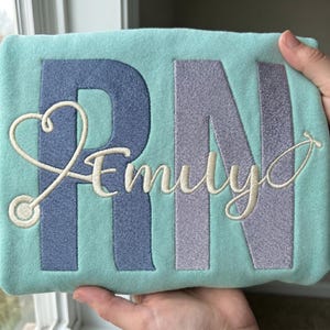 May include: A light teal sweatshirt with embroidered text and design. The large letters "RN" are in blue and lavender, with the name "Emily" in cursive. A stethoscope design forms a heart shape.