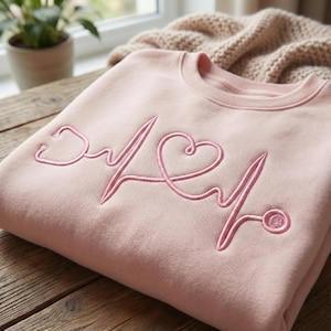 May include: A pale pink sweatshirt with a pink embroidered heartbeat design, including a stethoscope and heart. The garment is folded on a wooden surface, with a beige blanket visible in the background.