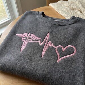 May include: Dark gray sweatshirt with a pink embroidered medical caduceus, heartbeat line, and heart design. The crew neck sweatshirt is made of a soft material and is suitable for medical professionals or those in the healthcare field.
