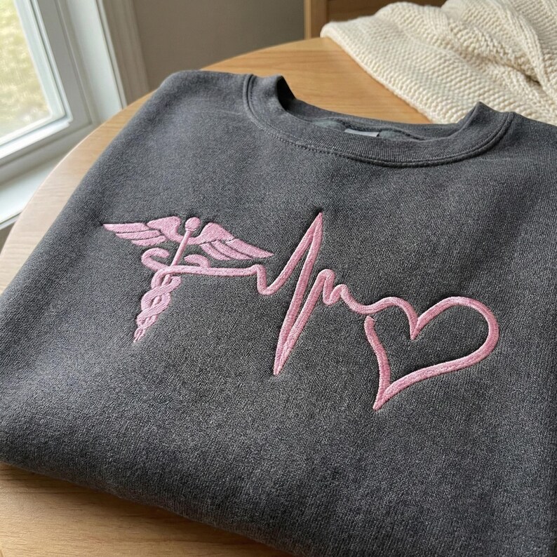 May include: Dark gray sweatshirt with a pink embroidered medical caduceus, heartbeat line, and heart design. The crew neck sweatshirt is made of a soft material and is suitable for medical professionals or those in the healthcare field.