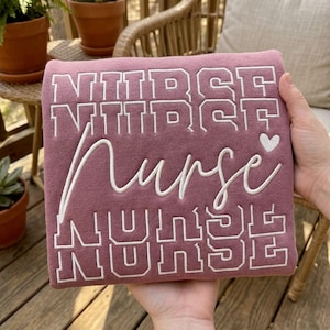 May include: A dusty rose sweatshirt featuring the word NURSE embroidered in a white block font across the top and bottom, with the word nurse in a white cursive script across the center, accompanied by a small white embroidered heart icon.