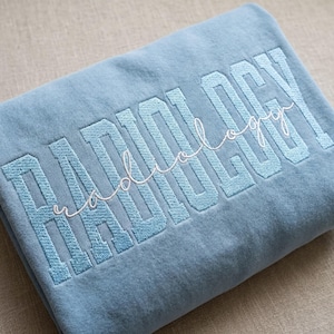 May include: A light blue sweatshirt with the word "RADIOLOGY" embroidered in a bold, light blue font. The word "radiology" is also embroidered in a cursive, white font over the top of the word "RADIOLOGY".