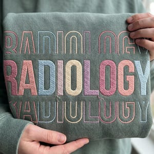 Puede incluir: Una sudadera verde salvia con la palabra RADIOLOGY bordada tres veces en un diseño apilado. Las letras están delineadas en varios colores pastel, incluyendo rosa, azul, amarillo y morado, creando un efecto de capas multicolor.