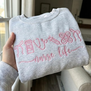 May include: A light gray sweatshirt with pink embroidery. The design includes a pill bottle, stethoscope, syringe, bandage, and bows, with the words "nurse life" in cursive. A cozy and stylish garment.