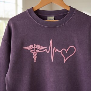 May include: A purple sweatshirt with a pink embroidered medical caduceus and heartbeat design. The crew neck sweatshirt is hanging on a wooden hanger. The design includes a heart shape.