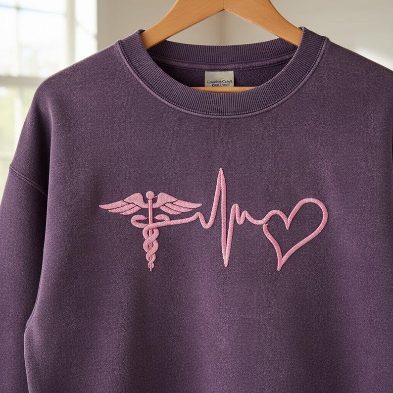 May include: A purple sweatshirt with a pink embroidered medical caduceus and heartbeat design. The crew neck sweatshirt is hanging on a wooden hanger. The design includes a heart shape.